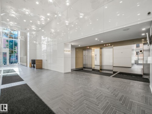 Spacious lobby featuring herringbone-patterned tile flooring, a high ceiling with intricate light fixtures, and twin stainless steel elevators - #1501 10180 103 St Nw, Edmonton, AB - Indoor Photo Showing Other Room