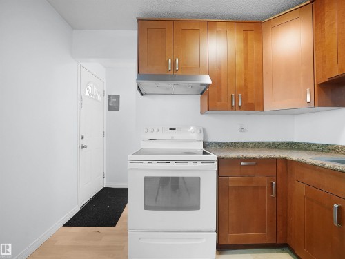 Compact kitchen featuring wood-finish cabinetry, speckled countertops, and a white electric range with an overhead stainless steel range hood - 10446 55 Avenue Nw, Edmonton, AB - Indoor Photo Showing Kitchen
