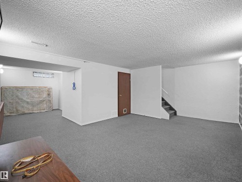 Spacious lower level featuring gray carpet flooring, white painted walls, a wood-finish door, and a partially finished ceiling with texture - 10446 55 Avenue Nw, Edmonton, AB - Indoor Photo Showing Other Room