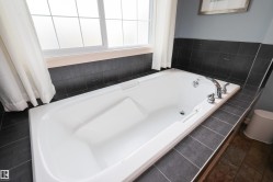 White soaking tub with chrome fixtures, surrounded by dark gray tile and a window with privacy glass - 