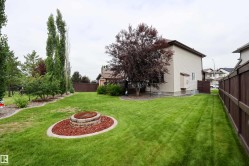 Expansive backyard featuring a lush lawn, a built-in stone fire pit with surrounding red rock, mature trees, and a privacy fence - 