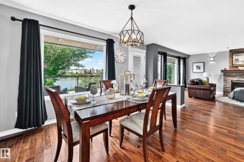 Dining area featuring wood-finish flooring, a contemporary chandelier, and a large window overlooking a body of water with distant structures - 12720 Hudson Way Nw, Edmonton, AB - Indoor Photo Showing Dining Room With Fireplace