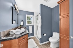 Bathroom featuring a wood-finish vanity with a dark countertop and backsplash, a framed mirror with contemporary lighting, a window with blinds, a toilet, and a tall wood-finish storage cabinet - 