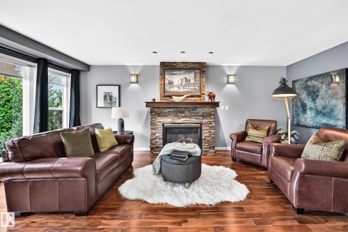 Living area featuring wood-finish flooring, a stacked stone fireplace with a wood mantel, and large windows - 12720 Hudson Way Nw, Edmonton, AB - Indoor Photo Showing Living Room With Fireplace