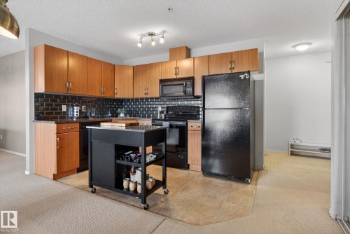 Kitchen featuring wood cabinetry, black subway tile backsplash, black appliances, and a movable island with a butcher block top - 423 2430 Guardian Road, Edmonton, AB - Indoor Photo Showing Kitchen