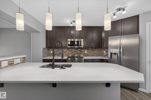 Kitchen island featuring a single-basin sink with a matte black faucet, pendant lighting, stainless steel appliances, dark wood cabinetry, and a mosaic tile backsplash - 3139 Carpenter Landing Sw, Edmonton, AB - Indoor Photo Showing Kitchen With Stainless Steel Kitchen With Double Sink With Upgraded Kitchen