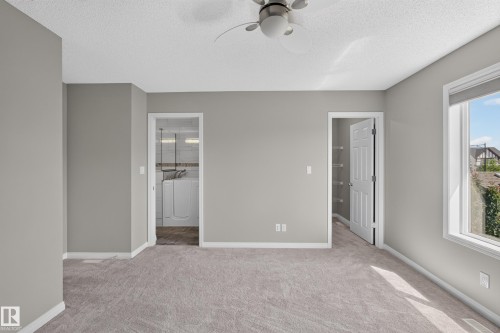 Spacious room featuring neutral carpeting, light gray wall tones, a ceiling fan, a window with a roller shade, and an adjacent laundry area with tiled flooring - 3139 Carpenter Landing Sw, Edmonton, AB - Indoor Photo Showing Other Room