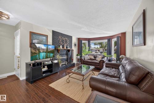 Spacious living area featuring wood-finish flooring, a contemporary black brick fireplace with a mantel, and a bay window with multiple panes - 8310 99 Avenue, Fort Saskatchewan, AB - Indoor Photo Showing Living Room With Fireplace