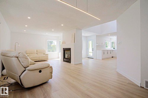 Spacious living area featuring light wood-finish flooring, a contemporary fireplace with a white surround, recessed lighting, a modern linear light fixture, and an abundance of natural light from the bay window - 11347 10 Avenue, Edmonton, AB - Indoor Photo Showing Living Room With Fireplace