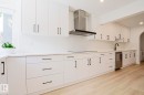 Modern kitchen featuring white cabinetry, a stainless steel dishwasher, a black wall-mounted range hood, light wood-finish flooring, and recessed lighting - 11347 10 Avenue, Edmonton, AB  - Indoor Photo Showing Kitchen 