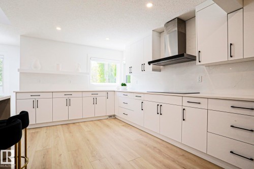 Bright kitchen featuring white cabinetry with matte black hardware, light wood-finish flooring, recessed lighting, and a contemporary stainless steel range hood - 11347 10 Avenue, Edmonton, AB - Indoor Photo Showing Kitchen