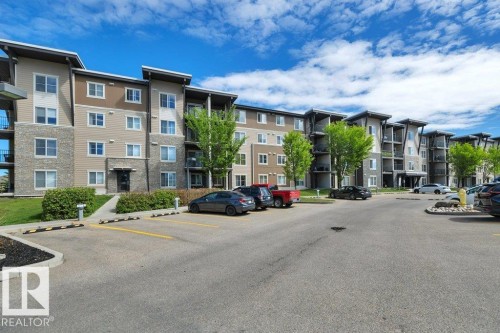Multi-story residential building featuring a stone and siding exterior, multiple balconies, and a paved parking area with designated spaces - #201 534 Watt Bv Sw, Edmonton, AB - Outdoor With Facade