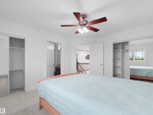Carpeted room featuring a ceiling fan with integrated lighting, two sliding closet doors (one mirrored), a doorway opening to a bathroom with a vanity, and an additional doorway revealing a stairwell with a wood banister - 3136 35 Avenue Nw, Edmonton, AB - Indoor Photo Showing Bedroom