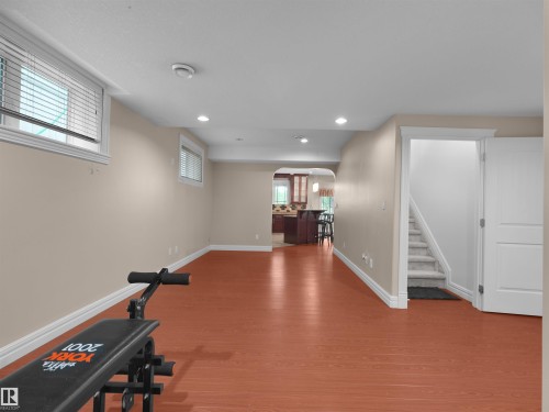 Spacious lower-level room featuring wood-finish flooring, two windows with blinds, recessed lighting, and direct access to a carpeted staircase and an arched entryway leading to a partial kitchen area - 3136 35 Avenue Nw, Edmonton, AB - Indoor
