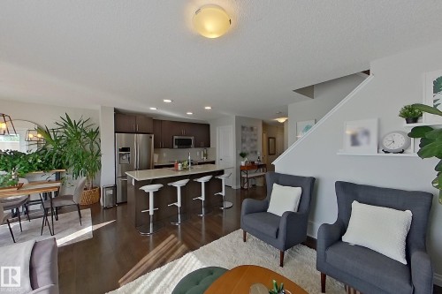 Open-concept living area featuring rich wood-finish flooring, a contemporary kitchen with dark cabinetry and stainless steel appliances, a kitchen island with seating, a dining area with modern lighting, and a staircase with white trim - 491 Ebbers Way, Edmonton, AB 