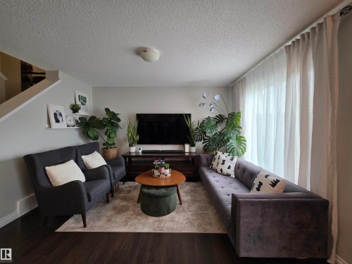 Spacious living area featuring dark wood-finish flooring, a neutral wall color, and a large window with sheer drapes - 491 Ebbers Way, Edmonton, AB 