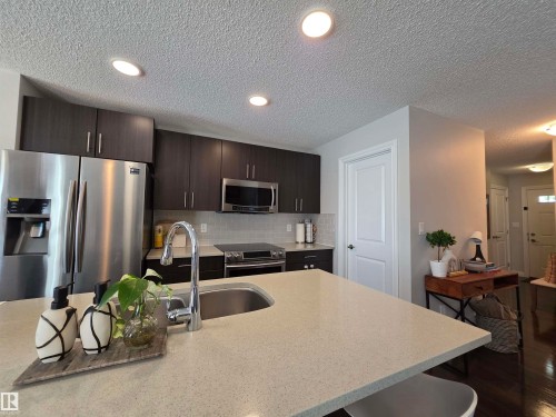 Kitchen featuring a light-toned countertop island with an undermount sink, dark wood-finish cabinetry, stainless steel appliances, and recessed lighting - 491 Ebbers Way, Edmonton, AB 