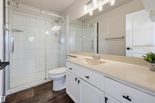 Full bathroom featuring a glass-enclosed shower with white subway tile, a white vanity with an integrated sink, a large mirror, and wood-finish flooring - 8716 130 Avenue Nw, Edmonton, AB - Indoor Photo Showing Bathroom