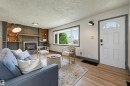 Living area featuring wood-finish flooring, a white paneled entry door with an arched transom, a large window, and a fireplace with a dark brick surround and wood-finish shelving - 8716 130 Avenue Nw, Edmonton, AB  - Indoor Photo Showing Living Room With Fireplace 