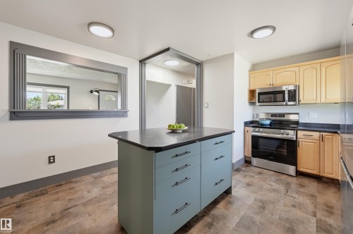 Kitchen featuring a central island with light blue cabinetry and dark countertop, wood-finish flooring, a pass-through window with decorative trim, stainless steel appliances, and light wood upper and lower cabinetry - 8716 130 Avenue Nw, Edmonton, AB - Indoor Photo Showing Kitchen