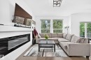 Modern living area featuring a linear electric fireplace with textured white tiling, a wood-finish flooring, and multiple large windows with natural light - 22270 99A Avenue Nw, Edmonton, AB  - Indoor Photo Showing Living Room With Fireplace 