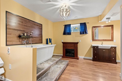 Bathroom featuring a freestanding white tub with a wood-panel accent wall, warm yellow paint, wood-finish flooring, a vanity with a rectangular sink, and a decorative fireplace - 1154 35A Avenue Nw, Edmonton, AB - Indoor With Fireplace