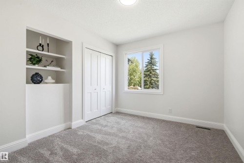Neutral-toned room featuring a window with exterior tree views, built-in shelving, white bi-fold closet doors, and plush gray carpet - 14335 47 Avenue Nw, Edmonton, AB - Indoor Photo Showing Other Room