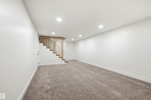 Spacious carpeted area featuring a natural wood-finish staircase with coordinating balusters, recessed ceiling lighting, crisp white walls, and matching baseboards - 14335 47 Avenue Nw, Edmonton, AB - Indoor Photo Showing Other Room