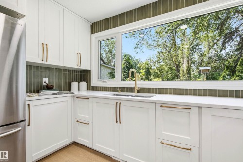 Modern kitchen featuring crisp white cabinetry, white countertops, a large picture window, a gold-toned gooseneck faucet, and a vertical tile backsplash - 14335 47 Avenue Nw, Edmonton, AB - Indoor Photo Showing Kitchen With Double Sink