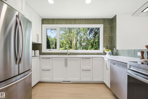 Kitchen featuring white shaker-style cabinetry, stainless steel appliances, a large window, a green vertical tile backsplash, and light wood-finish flooring - 14335 47 Avenue Nw, Edmonton, AB - Indoor Photo Showing Kitchen