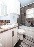 Bathroom featuring a white vanity with a stone-look countertop, a built-in tub with a shower, dark rectangular wall tiles with decorative accents, and overhead cabinetry - 