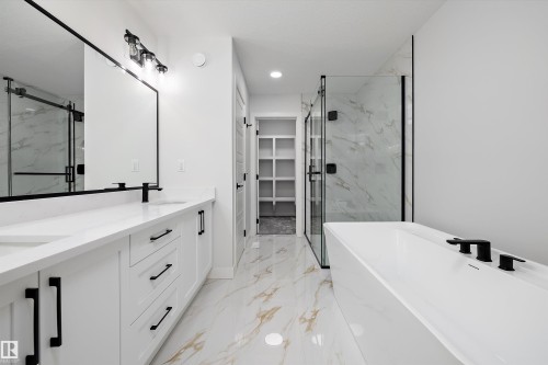 Modern bathroom featuring a dual vanity with white cabinetry and countertops, a freestanding soaking tub with matte black fixtures, a walk-in shower with clear glass enclosure and marble-patterned tile, and polished floor tiles with gold veining - 919 Elderberry Landing Nw, Edmonton, AB - Indoor Photo Showing Bathroom