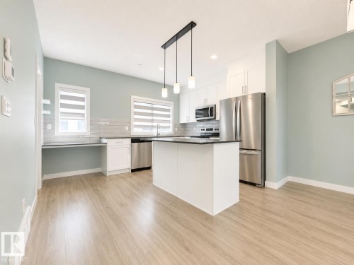 Open-concept kitchen featuring an island with a dark countertop, stainless steel appliances, white cabinetry, and wood-finish flooring - #56 804 Welsh Dr Sw, Edmonton, AB - Indoor Photo Showing Kitchen With Stainless Steel Kitchen With Upgraded Kitchen