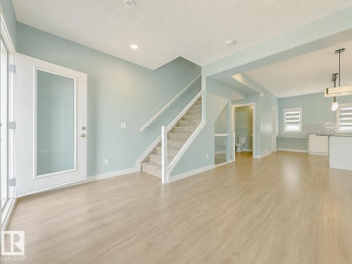 Open-concept living area featuring light wood-finish flooring, a contemporary staircase with carpeted treads, and a white framed glass-paneled exterior door - #56 804 Welsh Dr Sw, Edmonton, AB - Indoor