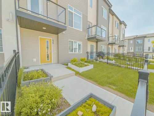 Modern exterior featuring a bright yellow entry door with vertical window panel, concrete walkway, raised garden beds, and a second-story balcony with glass railing - #56 804 Welsh Dr Sw, Edmonton, AB - Outdoor With Balcony