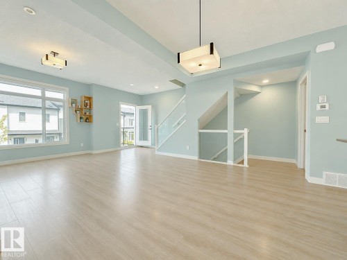Spacious main living area featuring light wood-finish flooring, abundant natural light from large windows and a glass door, and a contemporary ceiling light fixture - #56 804 Welsh Dr Sw, Edmonton, AB - Indoor Photo Showing Other Room