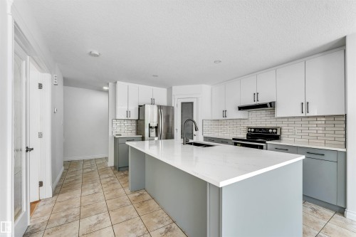 Modern kitchen featuring a large central island with a solid surface countertop, two-tone cabinetry with white upper and gray lower cabinets, full tile backsplash, stainless steel appliances, and tile flooring - 16205 55 Street Nw, Edmonton, AB - Indoor Photo Showing Kitchen With Upgraded Kitchen