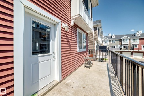 Private concrete balcony with black metal railing, featuring a white exterior door with glass insert, red horizontal siding, and an exterior wall sconce - #10 2803 James Mowatt Trail Sw, Edmonton, AB - Outdoor With Balcony