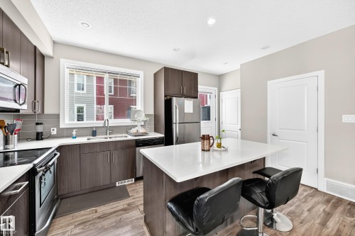 Modern kitchen featuring a central island with a solid surface countertop, wood-finish flooring, dark wood cabinetry, a grey subway tile backsplash, and stainless steel appliances - #10 2803 James Mowatt Trail Sw, Edmonton, AB - Indoor Photo Showing Kitchen