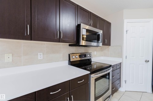 Kitchen featuring dark wood-finish cabinetry, light solid-surface countertops, stainless steel appliances, a tiled backsplash, and tiled flooring - 1919 Hammond Place, Edmonton, AB - Indoor Photo Showing Kitchen With Upgraded Kitchen