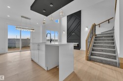 Open concept living area featuring wood-finish flooring, a contemporary kitchen island with a white countertop and dark faucet, a modern staircase with wood and metal railing, a striking black accent wall, and a sliding glass door leading to the exterior - 