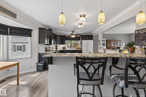 Open-concept kitchen featuring an eat-at island with a light-toned granite countertop, dark wood-finish cabinetry, and a tile backsplash - 10 Abbey Crescent, St. Albert, AB - Indoor Photo Showing Kitchen With Upgraded Kitchen