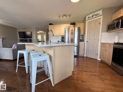 Kitchen featuring wood-finish flooring, light wood cabinetry, stainless steel appliances, a white subway tile backsplash, and a breakfast bar with a light-toned countertop - 1820 Robertson Crescent Sw, Edmonton, AB - Indoor Photo Showing Kitchen