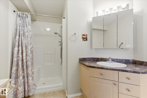 Bathroom featuring a white shower stall with an adjustable showerhead, a light wood-finish vanity with a dark countertop, an integrated sink, and a triple mirrored medicine cabinet with overhead lighting - #230 8802 Southfort Dr, Fort Saskatchewan, AB - Indoor Photo Showing Bathroom