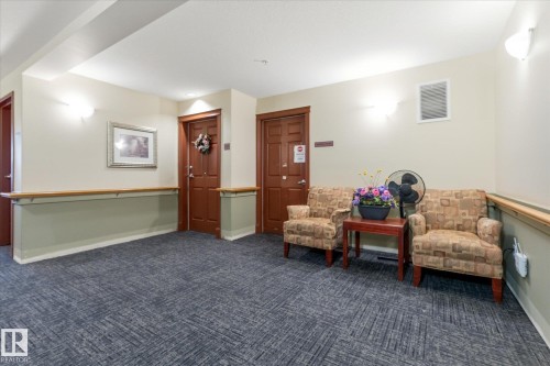 Carpeted common area featuring sconce lighting, natural wood handrails, and wood-finish doors - #230 8802 Southfort Dr, Fort Saskatchewan, AB - Indoor Photo Showing Other Room