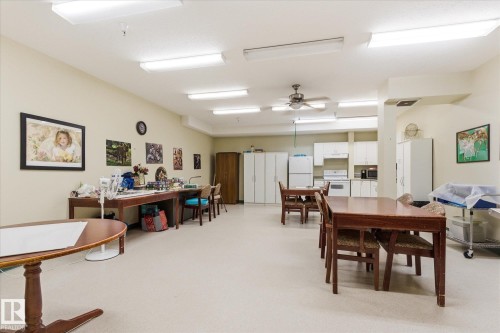 Open-concept common area featuring recessed fluorescent lighting, light-toned flooring, a built-in kitchen area with white cabinetry, and multiple activity tables - #230 8802 Southfort Dr, Fort Saskatchewan, AB - Indoor Photo Showing Other Room