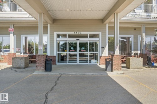 Entryway featuring automatic glass sliding doors, a covered portico, brick accents, and light-colored siding - #230 8802 Southfort Dr, Fort Saskatchewan, AB - Outdoor