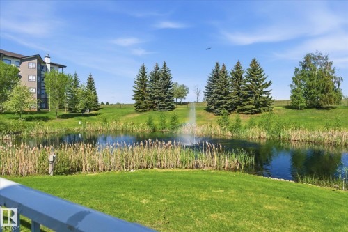 Serene pond with a central fountain, surrounded by lush green grass, mature trees, and natural reeds - #230 8802 Southfort Dr, Fort Saskatchewan, AB - Outdoor With Body Of Water With View