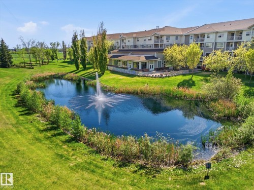 Tranquil pond with an active fountain feature, surrounded by lush green landscaping and mature trees - #230 8802 Southfort Dr, Fort Saskatchewan, AB - Outdoor With Body Of Water With View