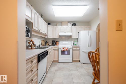Kitchen featuring L-shaped cabinetry, light-toned countertops, tile flooring, and white appliances including a refrigerator, range, and dishwasher - #310 17151 94A Av Nw, Edmonton, AB - Indoor Photo Showing Kitchen With Double Sink
